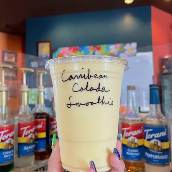 A hand holds a Caribbean Colada Smoothie from Grouchy John's Coffee, with syrup bottles in the background.