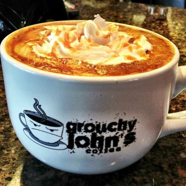A Grouchy John's Coffee mug filled with a caramel latte, topped with whipped cream and caramel drizzle.