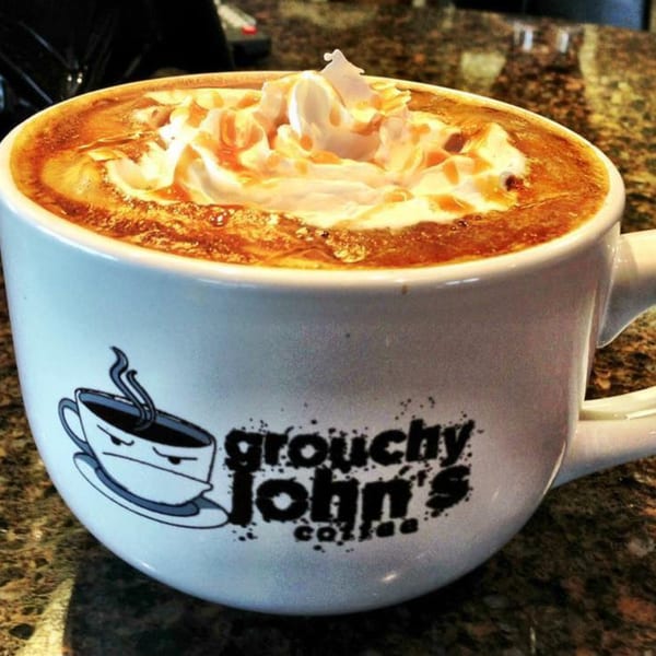A Grouchy John's Coffee mug filled with a caramel latte topped with whipped cream and drizzle.