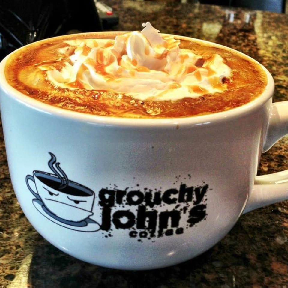 A Grouchy John's Coffee mug filled with a caramel latte topped with whipped cream and drizzle.